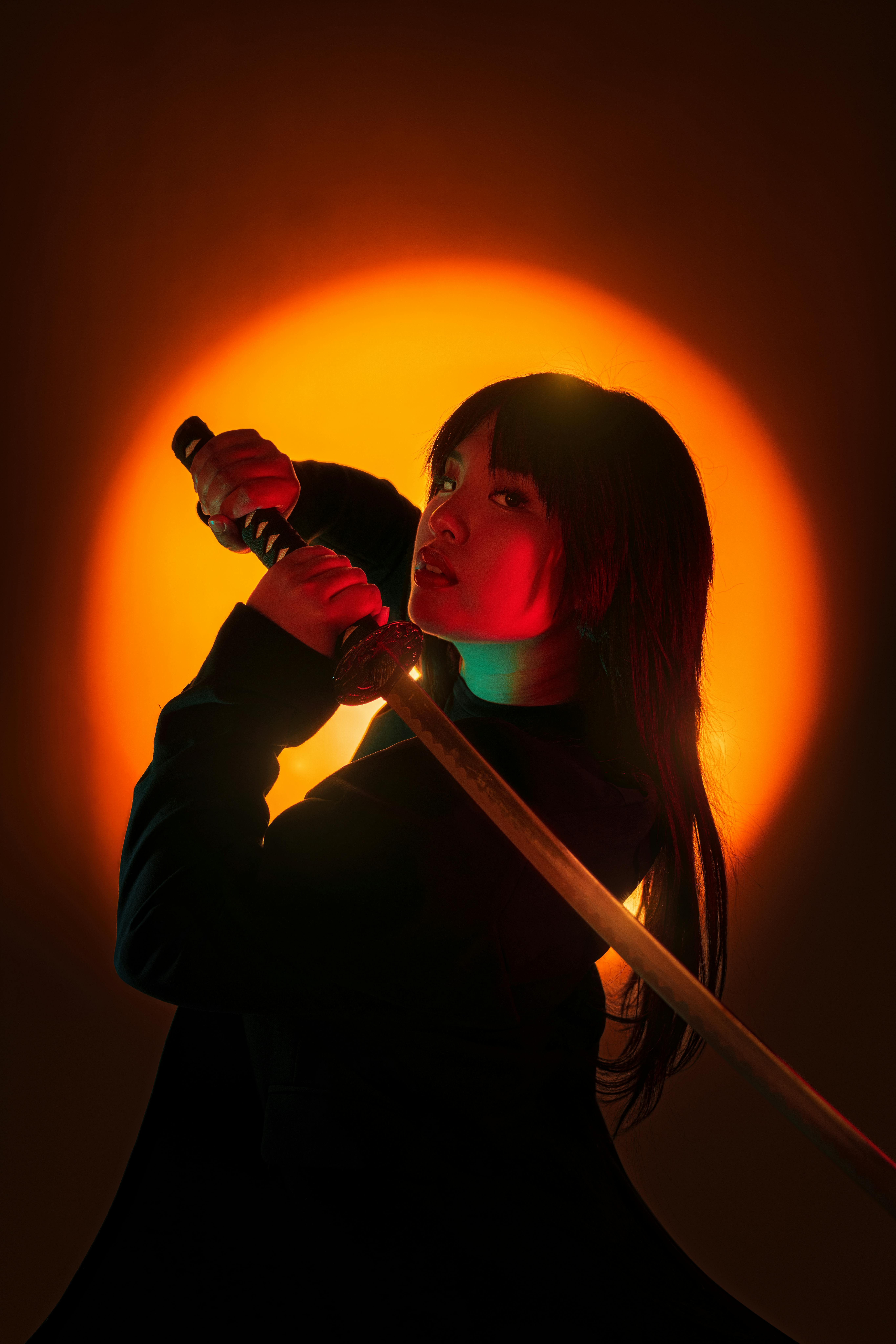 A person holding a katana sword, posed against a bright orange circular background, creating a dramatic silhouette effect.