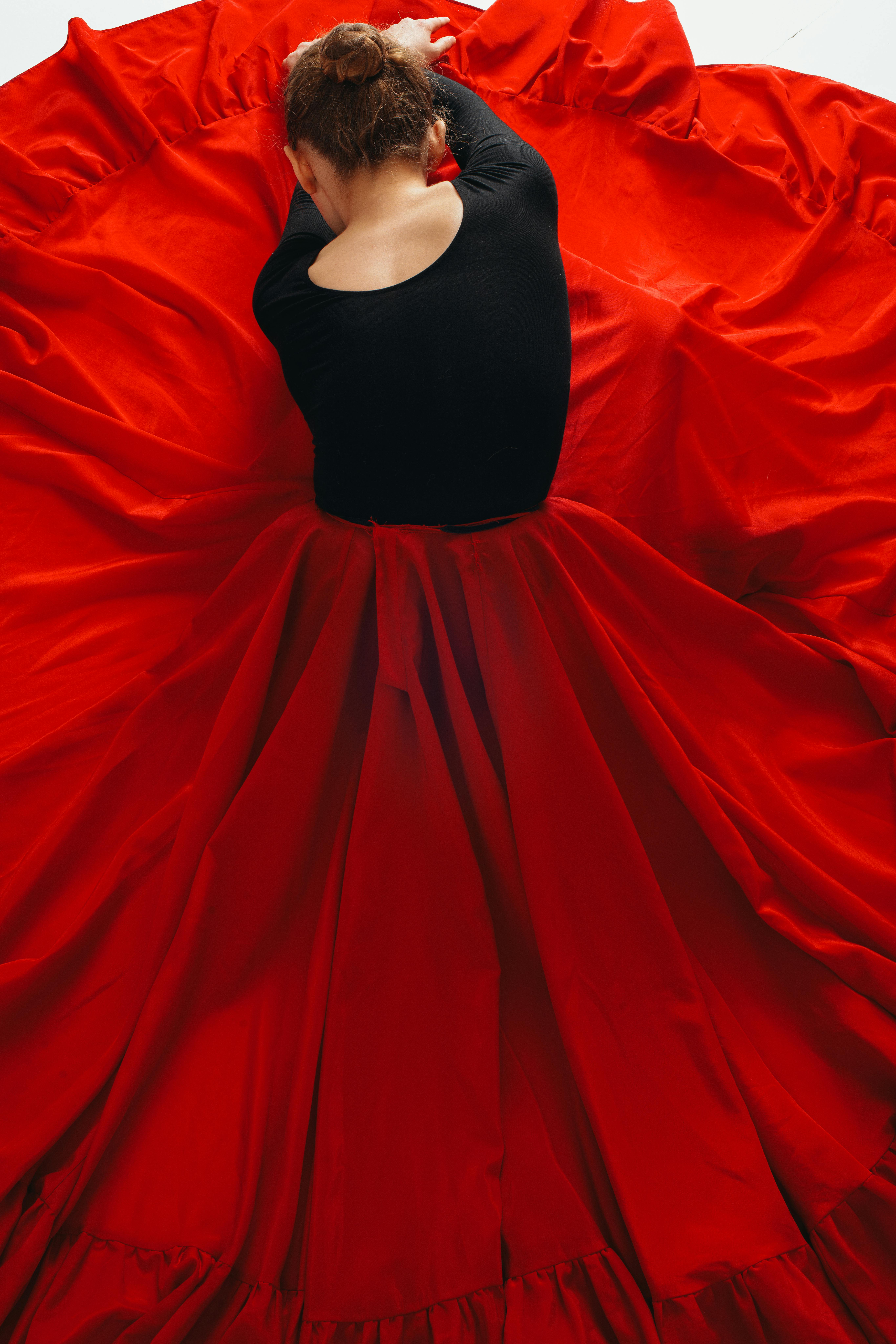 A person with their back turned is seated, wearing a black top and surrounded by a large, flowing red skirt that creates a dramatic visual effect.