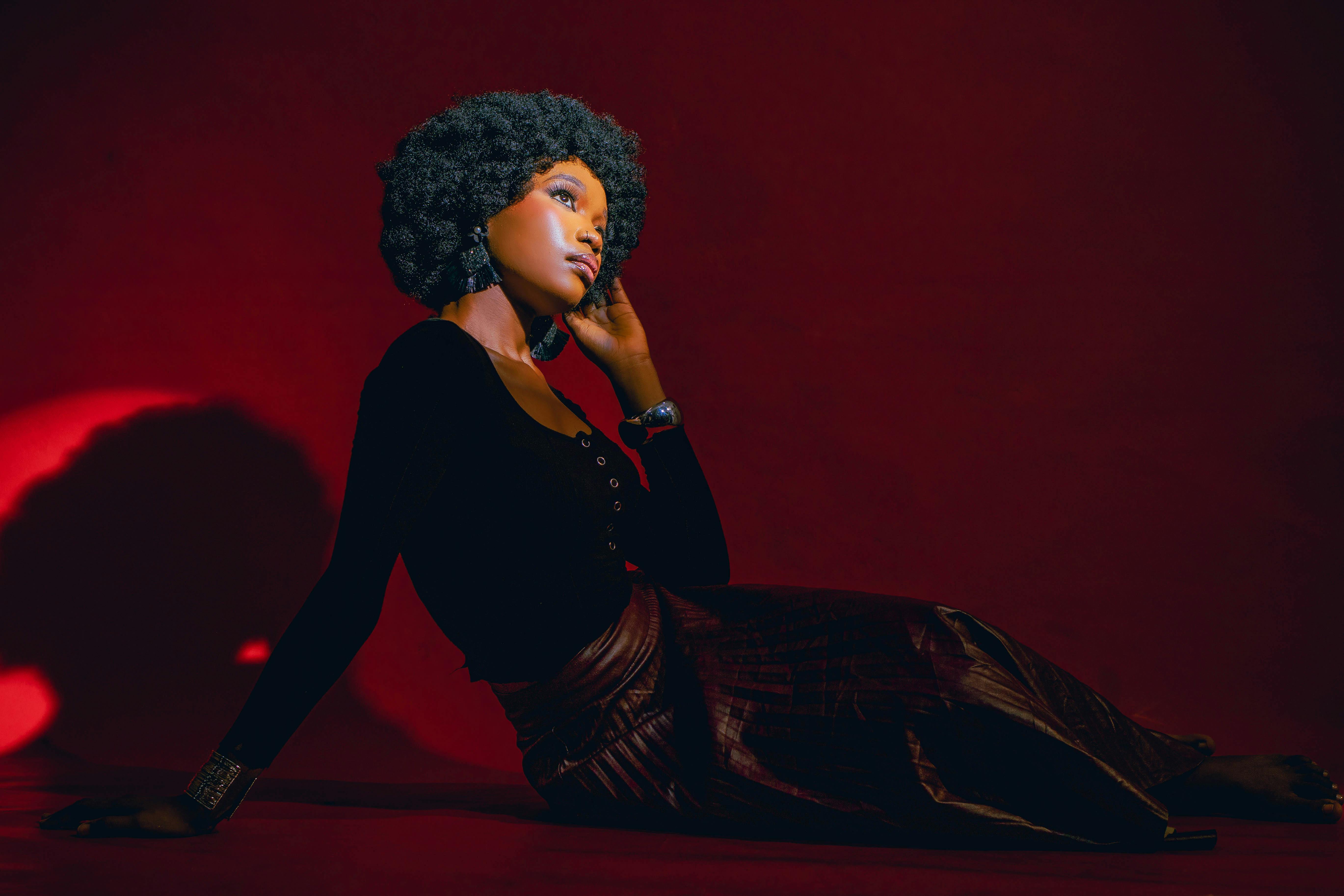 A person with a textured hairstyle sitting against a dark red background, wearing a black top and a long, pleated skirt, with earrings that catch the light and a contemplative expression.